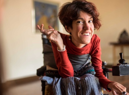 Woman sitting in a wheelchair, smiling at something off camera, gesturing.