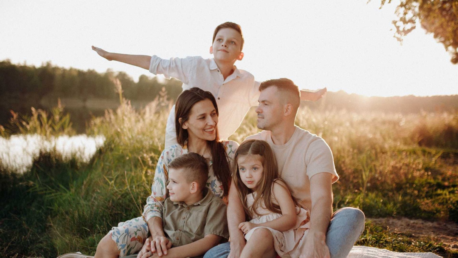 Family sitting together outside as the sun is setting. One child holding arms out like an aeroplane, the other two children sitting on their parents' laps.