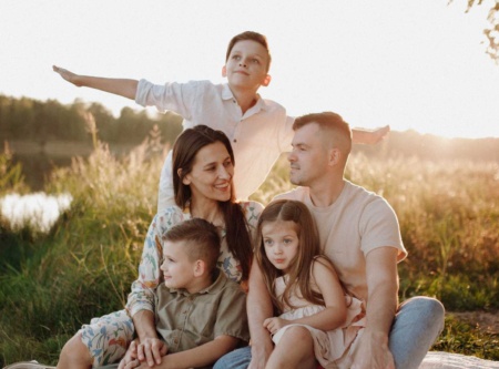 Family sitting together outside as the sun is setting. One child holding arms out like an aeroplane, the other two children sitting on their parents' laps.