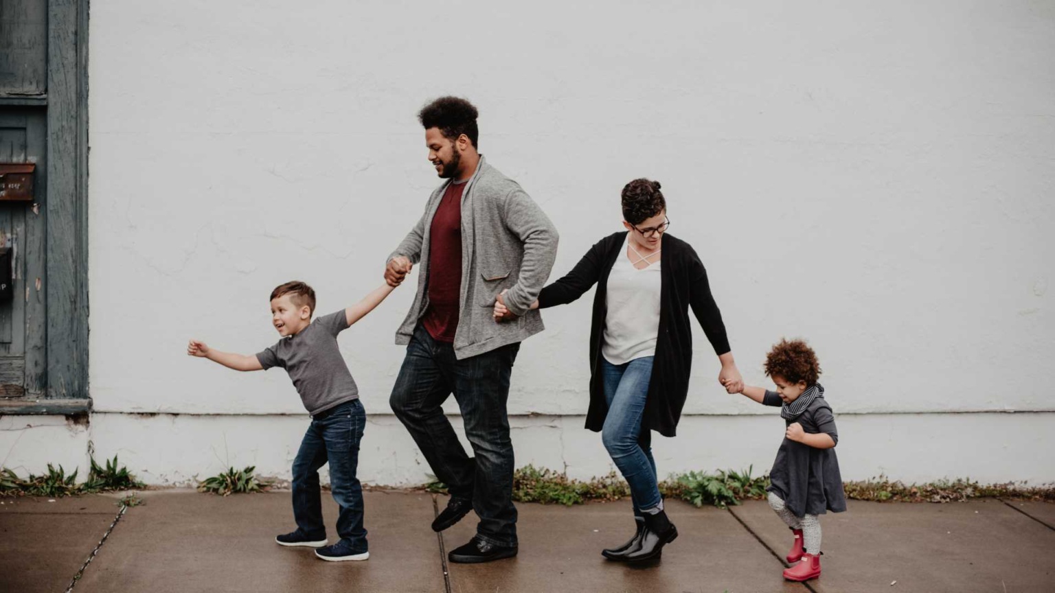 Young family walking along the footpath, holding hands. Young son leading and holding Dad's hand, who's hold Mum's hand. Mum is looking backwards to her young daughter, also holding hands.
