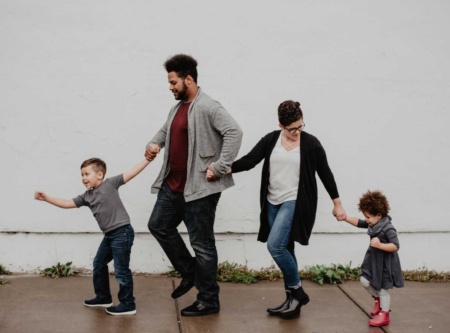 Young family walking along the footpath, holding hands. Young son leading and holding Dad's hand, who's hold Mum's hand. Mum is looking backwards to her young daughter, also holding hands.